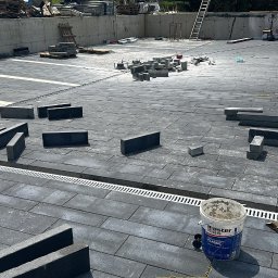 Uslugi ogolnobudowlane - Układanie ciemnoszarej kostki brukowej z jasnymi pasami w Rajcza, widoczne korytka odwadniające i paleta z kostką w tle.