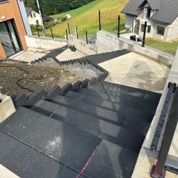 Uslugi ogolnobudowlane - Schody zewnętrzne wyłożone czarną kostką brukową, z betonowymi ścianami oporowymi i metalowymi barierkami, w tle dom w budowie i zielony krajobraz górski.