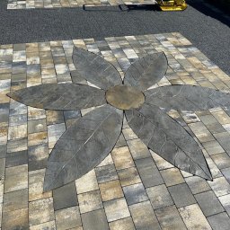 Uslugi ogolnobudowlane - Ułożona kostka brukowa w kształcie kwiatu z szarymi liśćmi na tle mozaiki z kostek w odcieniach szarości i beżu, obok pas z drobnego szarego kruszywa.