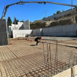 Wylewka fundamentowa zbrojona siatką stalową, pracownik w czerwonej czapce i żółtych rękawicach przy pracy, w tle betonowy mur oporowy i ramię pompy do betonu.