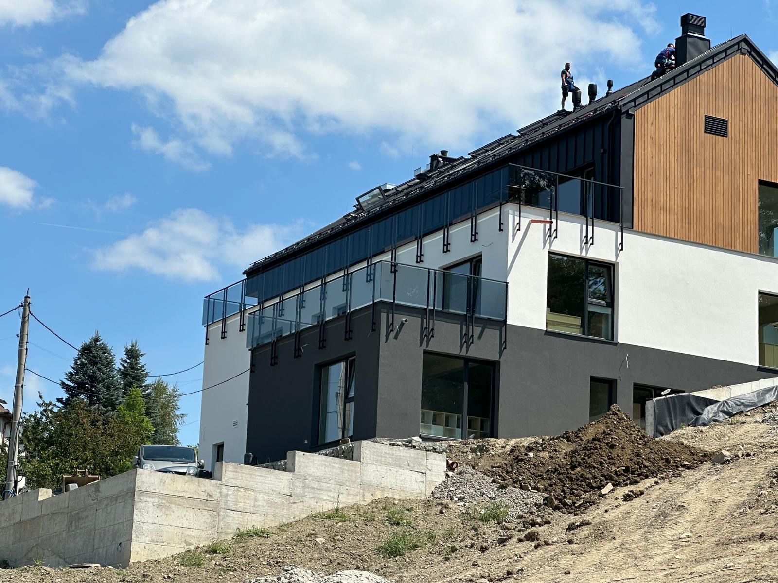 Dwóch pracowników na dachu nowoczesnego domu z drewnianą elewacją, widok z dołu na tle błękitnego nieba z chmurami, teren przed domem w trakcie niwelacji.