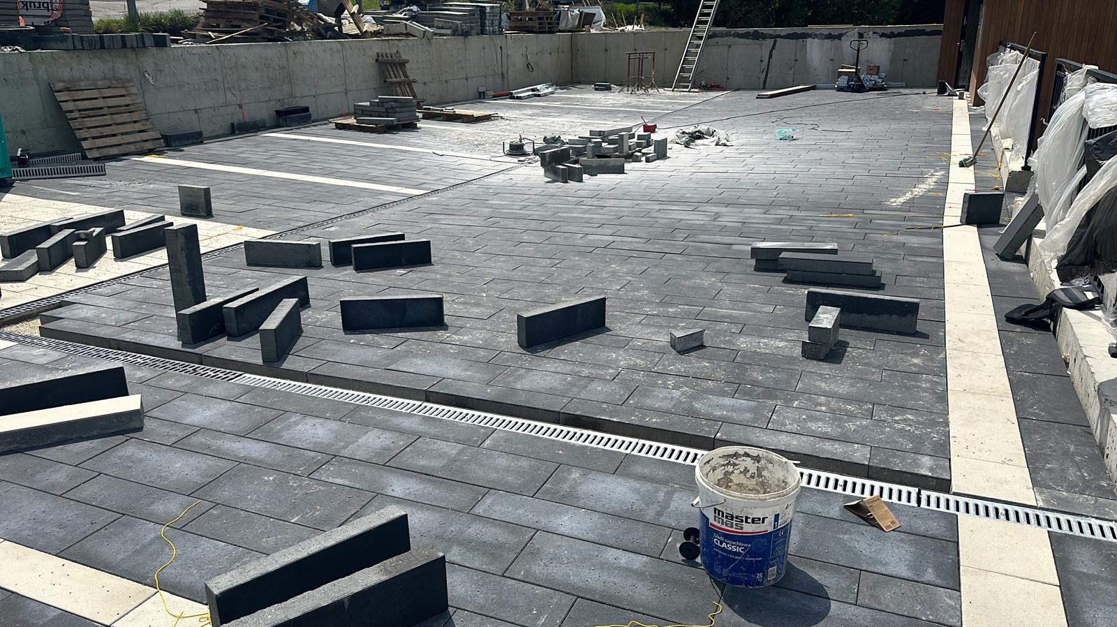 Układanie ciemnoszarej kostki brukowej z jasnymi pasami w Rajcza, widoczne korytka odwadniające i paleta z kostką w tle.