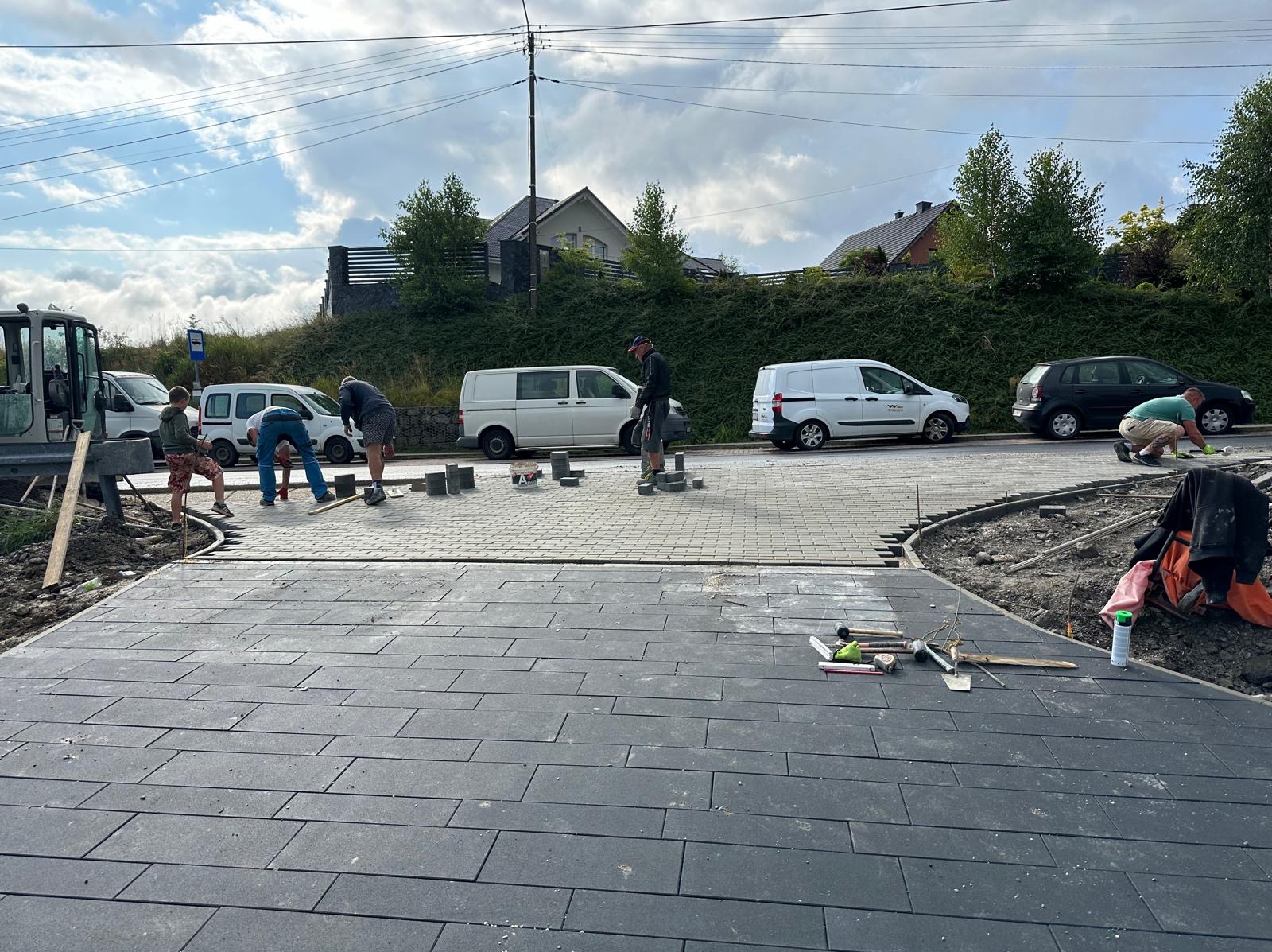 Ekipa brukarzy układa ciemnoszarą kostkę brukową na podjeździe, widoczne narzędzia pracy i zaparkowane samochody w tle, częściowo ułożona kostka w dwóch odcieniach szarości wyznacza zakrzywiony...