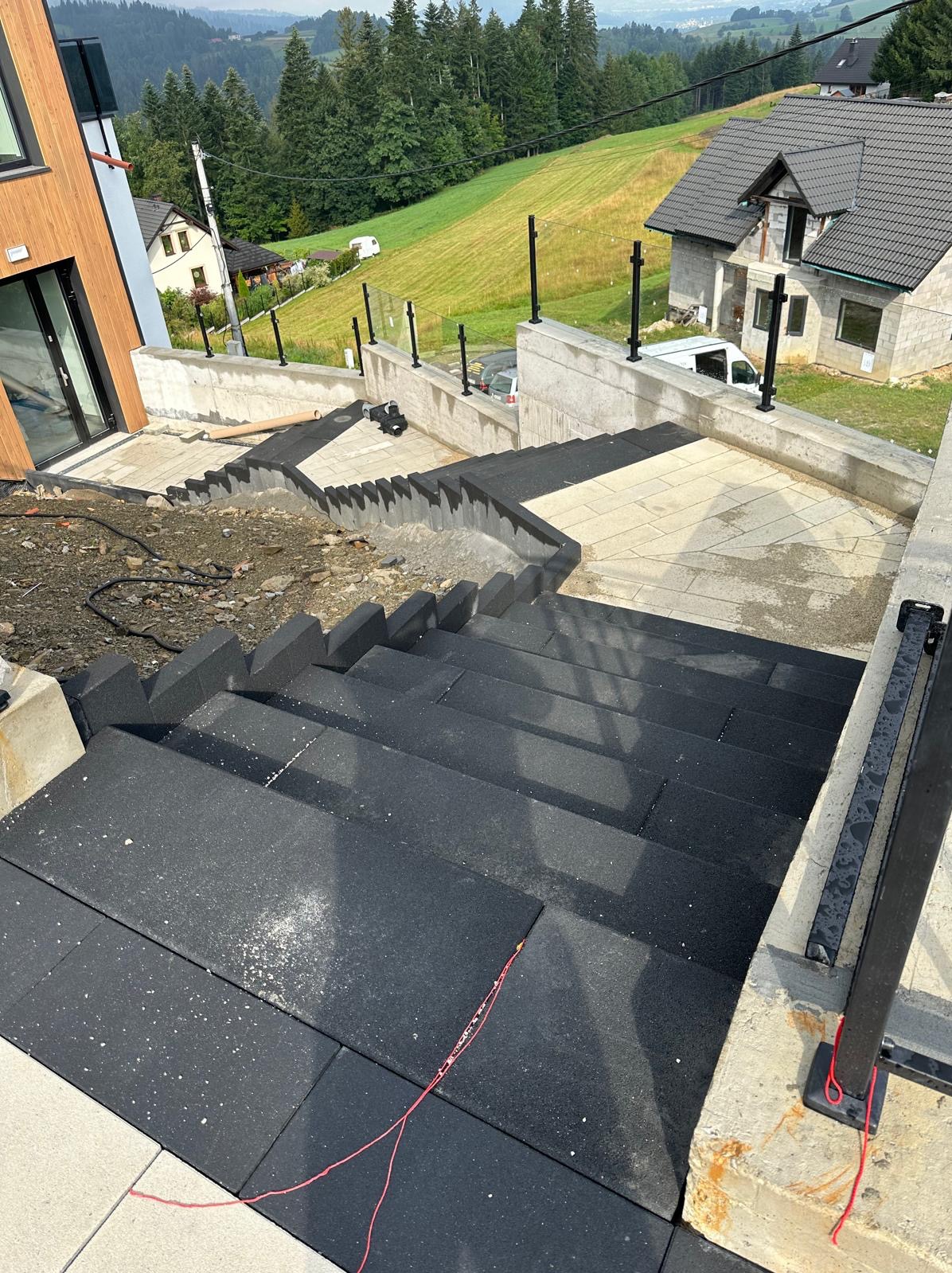 Schody zewnętrzne wyłożone czarną kostką brukową, z betonowymi ścianami oporowymi i metalowymi barierkami, w tle dom w budowie i zielony krajobraz górski.