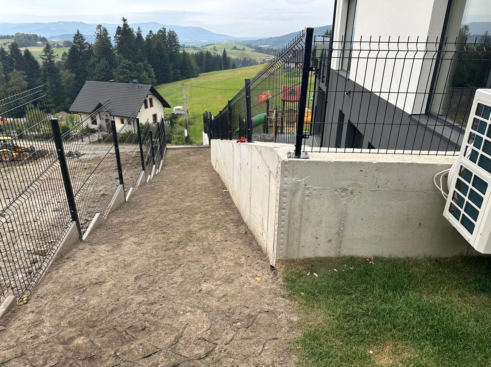 Ukształtowany teren działki z widocznym ogrodzeniem panelowym na betonowej podmurówce, w tle dom z klimatyzatorem i plac zabaw, a dalej rozległy krajobraz zalesionych wzgórz.