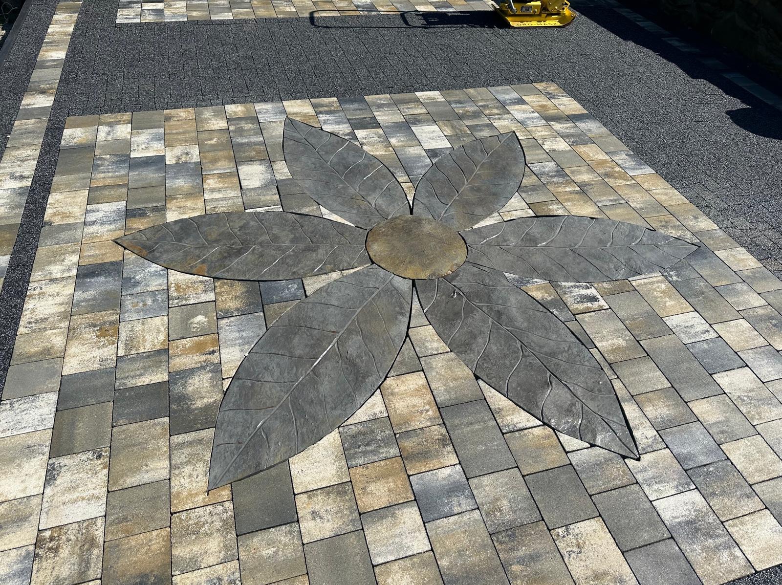 Ułożona kostka brukowa w kształcie kwiatu z szarymi liśćmi na tle mozaiki z kostek w odcieniach szarości i beżu, obok pas z drobnego szarego kruszywa.