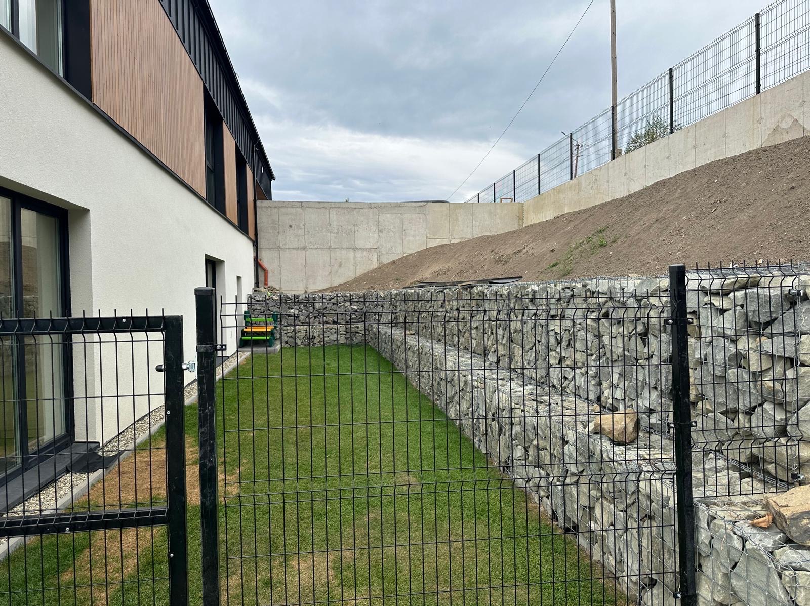 Gabionowe mury oporowe z czarnej siatki, zabezpieczające skarpę ziemną za budynkiem z elewacją w kolorze białym i drewnianym brązie, widoczne elementy systemu odwadniającego i zielony trawnik.