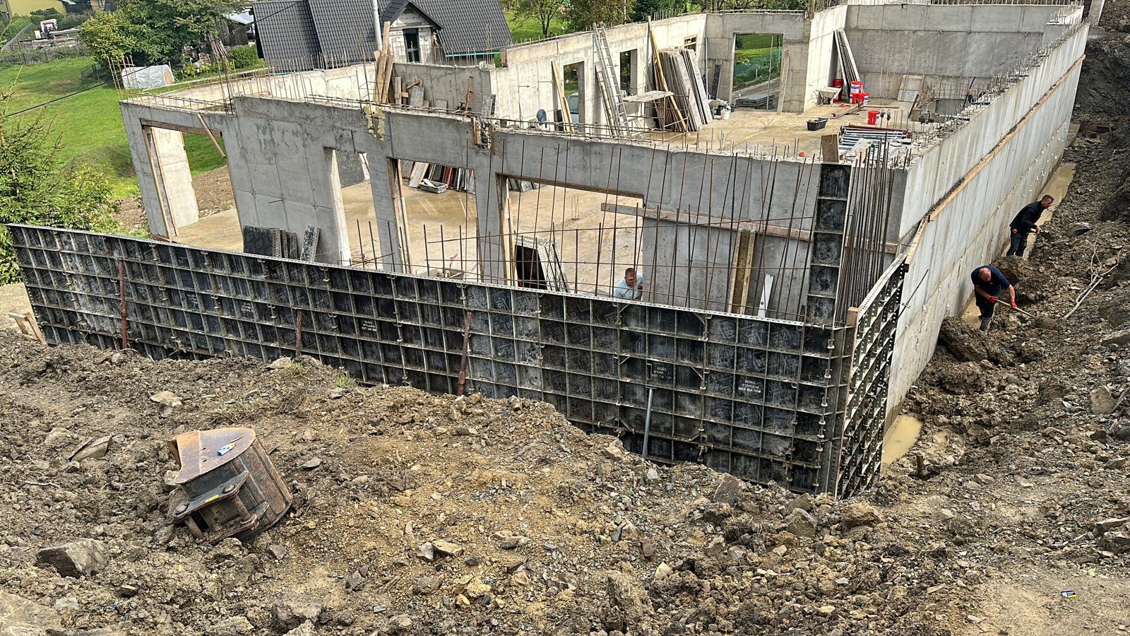 Budowa domu z betonu komórkowego w trakcie realizacji: widoczne szalunki, zbrojenia, fundamenty i pracujący robotnicy przy wyrównywaniu terenu wokół budynku.