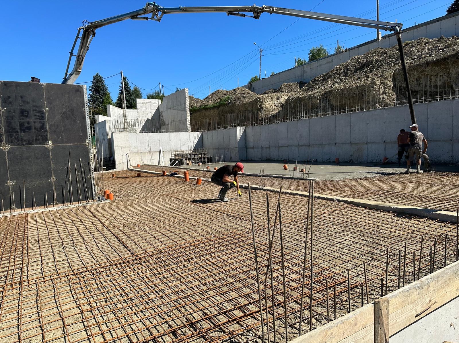 Wylewka fundamentowa zbrojona siatką stalową, pracownik w czerwonej czapce i żółtych rękawicach przy pracy, w tle betonowy mur oporowy i ramię pompy do betonu.