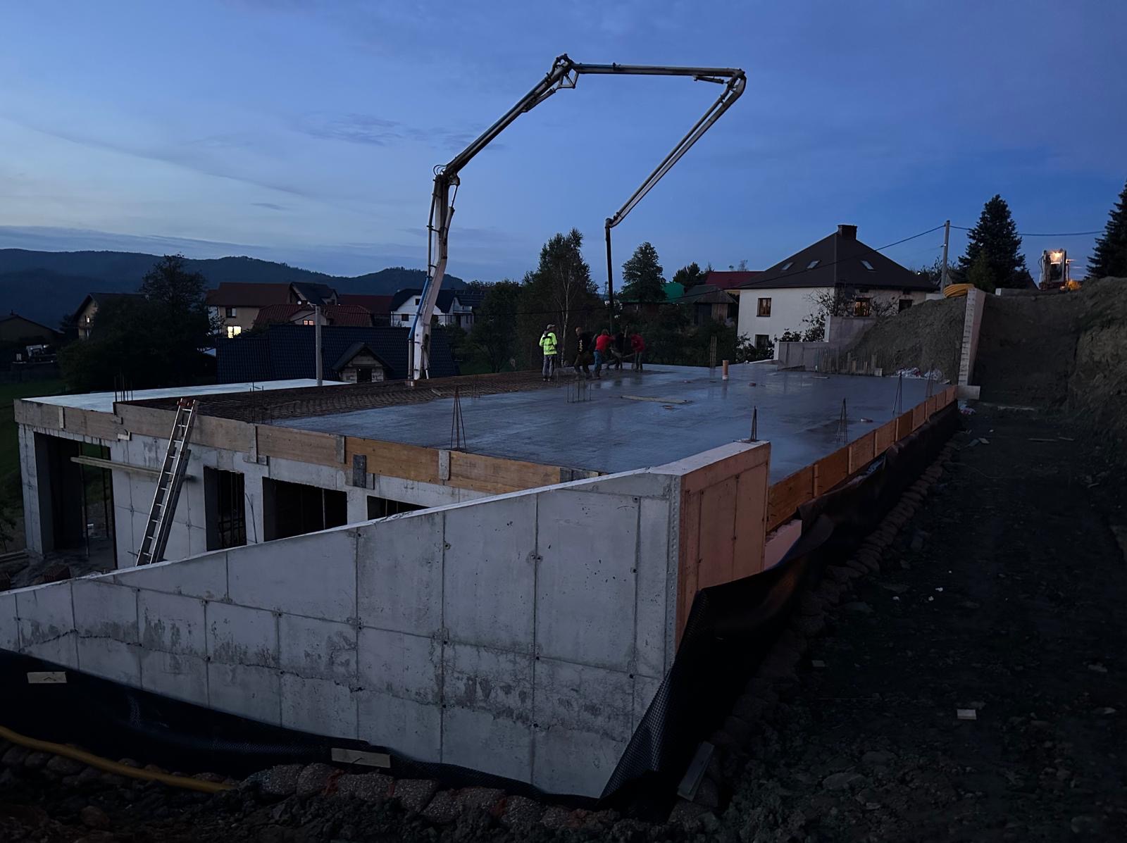 Wylewanie betonu na strop domu murowanego w trakcie budowy, widoczna pompa do betonu i grupa pracowników w odblaskowych kamizelkach, tło stanowią domy jednorodzinne i wzgórza w porannym świetle.