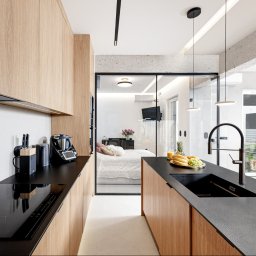PBE - Nowoczesna kuchnia z drewnianymi frontami i czarnym blatem, widok na sypialnię przez szklane drzwi. Minimalistyczny design, oświetlenie punktowe, owocowy poczęstunek na blacie.