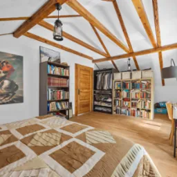 Jasna sypialnia na poddaszu z drewnianymi belkami, łóżkiem z patchworkową narzutą i biurkiem. Obraz Napoleona na ścianie dodaje charakteru.