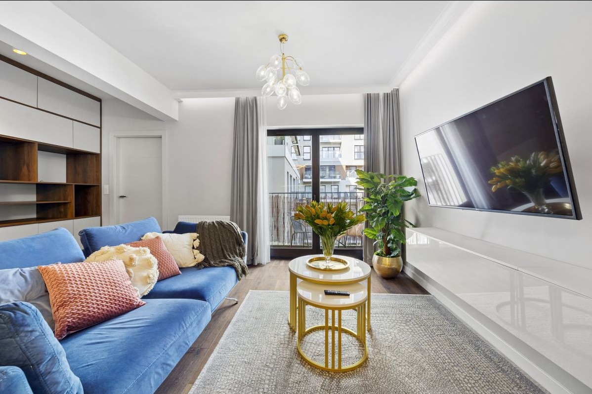 Jasny salon z niebieską sofą, okrągłymi stolikami kawowymi z pozłacanymi nogami, telewizorem na ścianie i balkonem z widokiem na budynek mieszkalny.