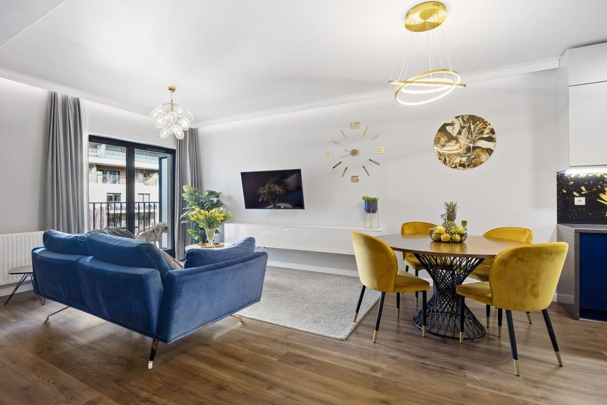 Jasne wnętrze mieszkania z granatową sofą, okrągłym stołem z żółtymi krzesłami, dekoracjami na ścianie i parkietem