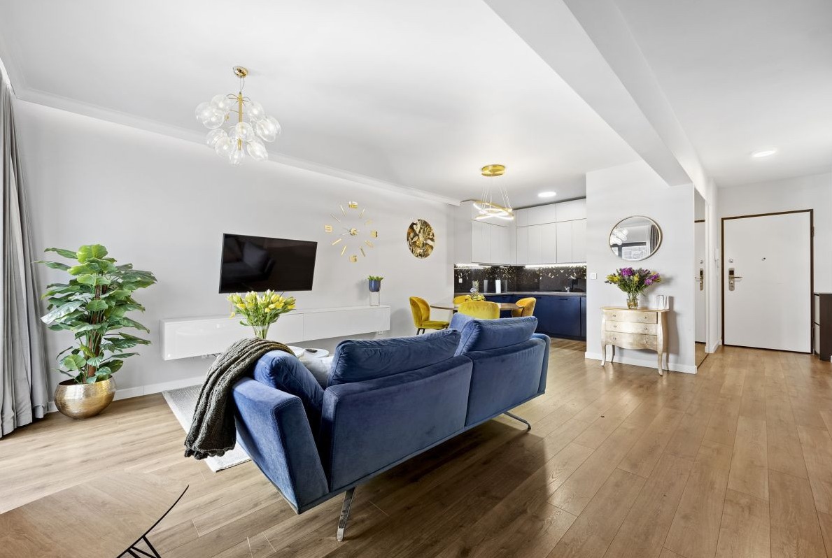 Nowoczesne wnętrze mieszkania z niebieską sofą, drewnianą podłogą, otwartą kuchnią z granatowymi szafkami i złotymi akcentami, oświetlone dekoracyjnymi lampami.