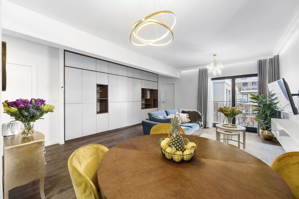 Jasne, nowocześnie urządzone wnętrze mieszkania z drewnianym stołem, żółtymi krzesłami, niebieską sofą, zabudową ścienną z białymi szafkami i dekoracyjnym oświetleniem sufitowym.