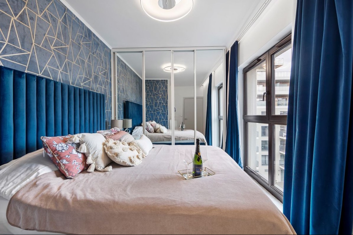 Elegancka sypialnia z niebieskim, geometrycznym wzorem na ścianie, welurowym zagłówkiem i butelką szampana na łóżku, odbicie w lustrzanych drzwiach szafy.