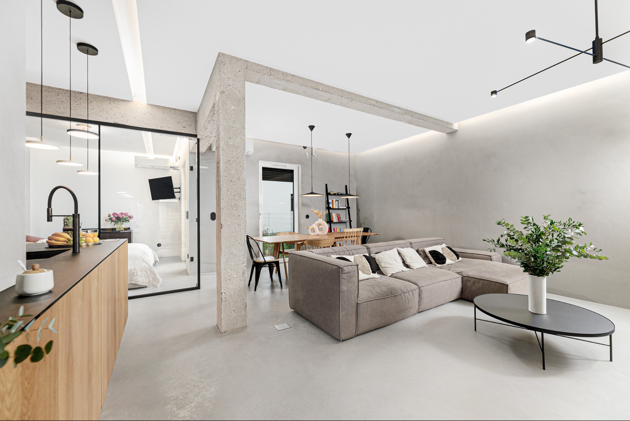 Nowoczesne wnętrze: salon z sofą narożną, jadalnia z drewnianym stołem i kuchnia z czarnym zlewem. Widok na sypialnię przez szklaną ścianę. Minimalistyczny design.