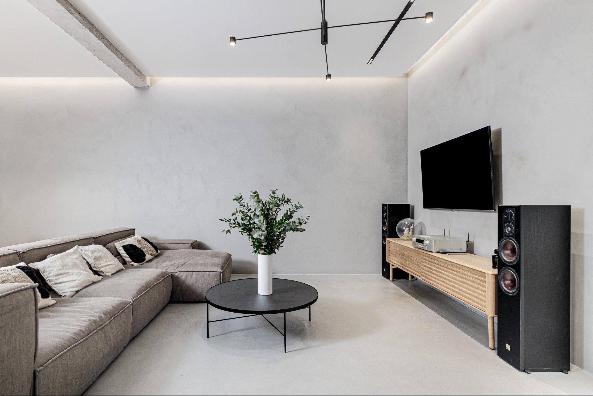 Jasne, minimalistyczne wnętrze salonu z szarym narożnikiem, okrągłym stolikiem kawowym i szafką RTV z telewizorem. Na suficie nowoczesna lampa.