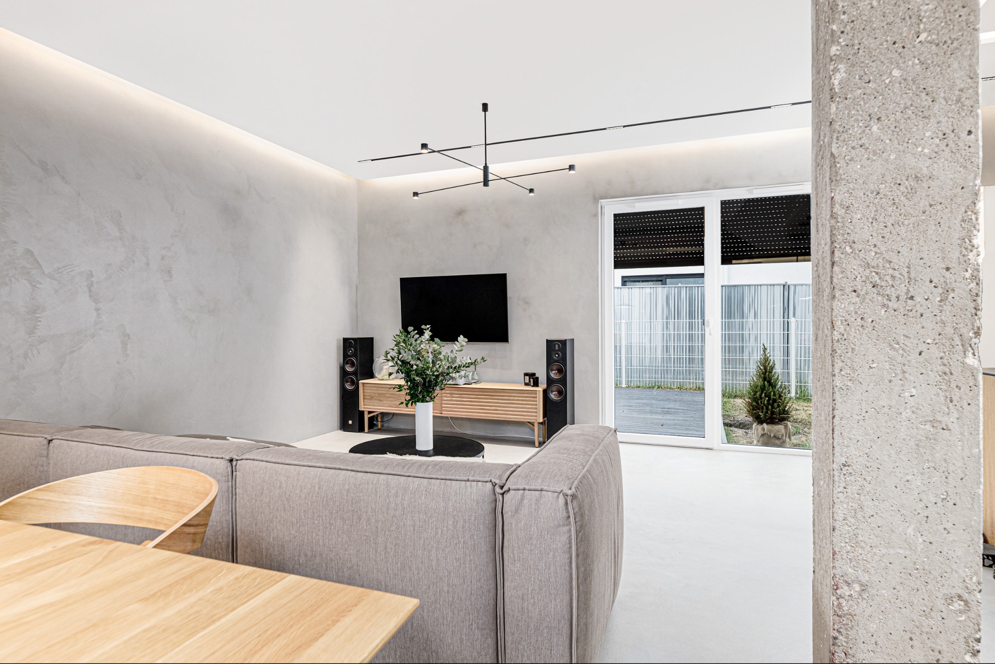 Nowoczesny salon z szarą ścianą dekoracyjną, jasną podłogą i minimalistycznym oświetleniem. Widoczny fragment sofy i stołu. Kolumna betonowa w prawym rogu.