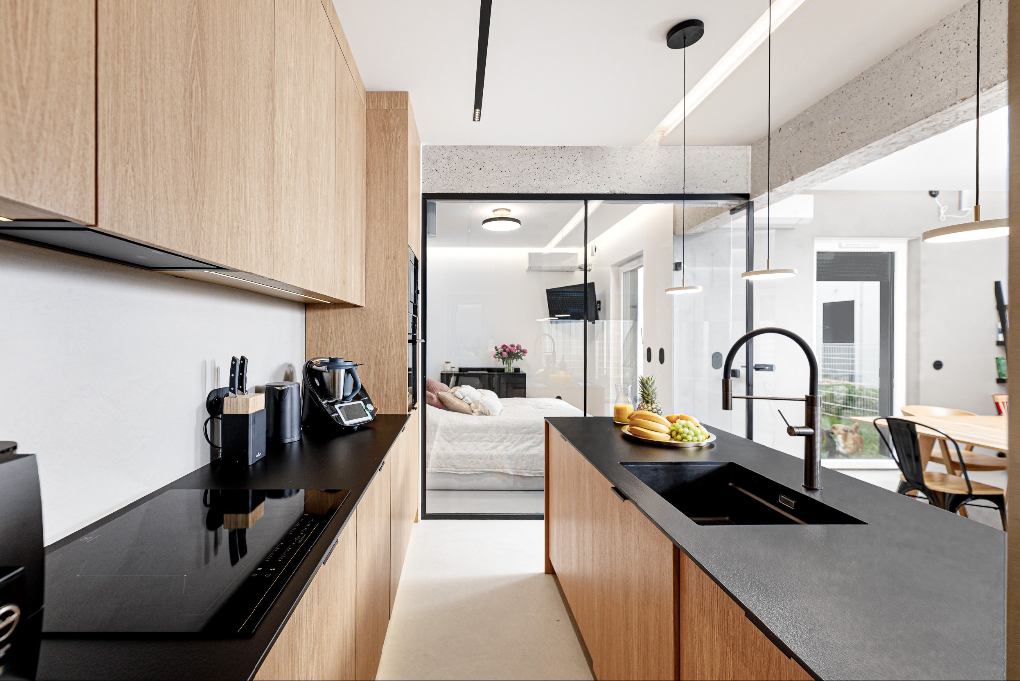 Nowoczesna kuchnia z drewnianymi frontami i czarnym blatem, widok na sypialnię przez szklane drzwi. Minimalistyczny design, oświetlenie punktowe, owocowy poczęstunek na blacie.