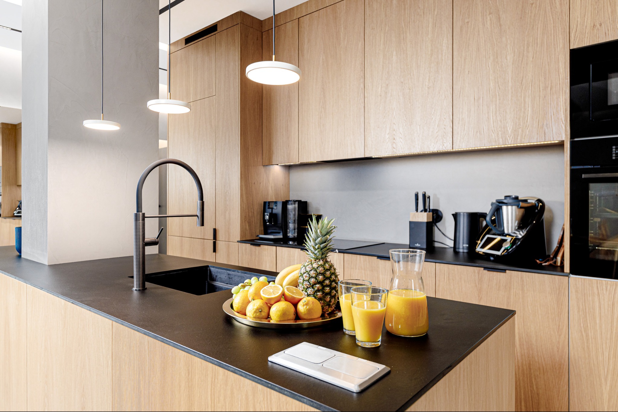 Jasna kuchnia z drewnianymi frontami, czarnym blatem i owocami na wyspie. Nowoczesne oświetlenie punktowe i sprzęt AGD w tle. Minimalistyczny design.