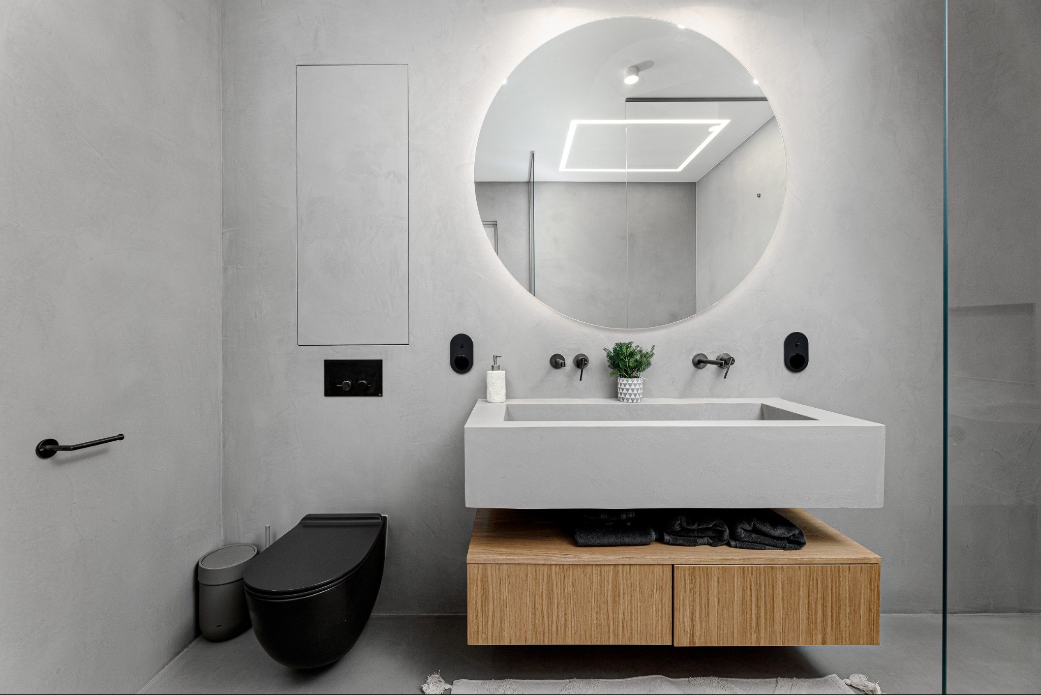 Minimalistyczna łazienka z betonową umywalką, okrągłym lustrem z podświetleniem LED i czarną toaletą. Szare ściany z fakturą betonu. Nowoczesny design.