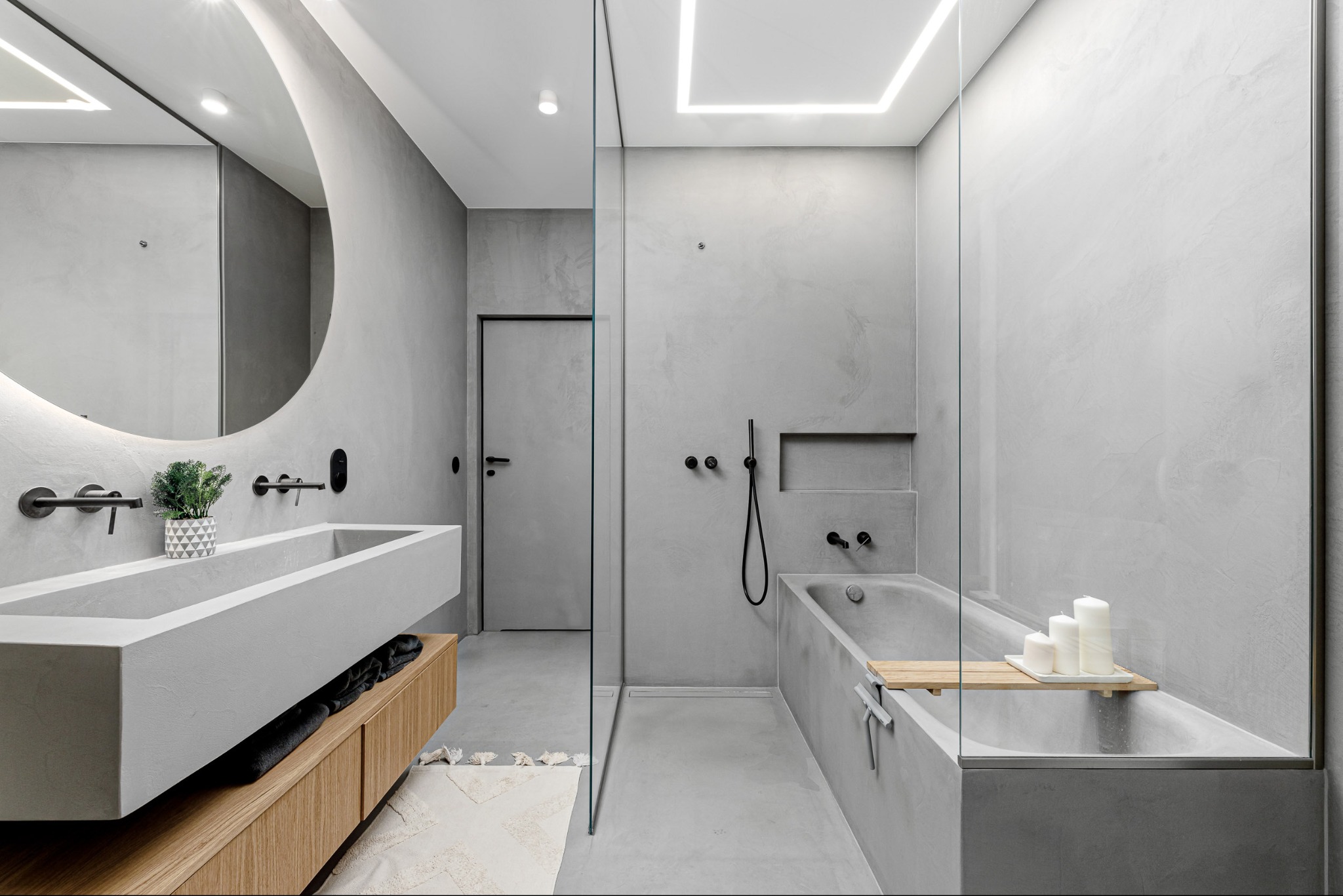 Nowoczesna łazienka w odcieniach szarości z betonową umywalką, okrągłym lustrem z podświetleniem i wanną za szklaną ścianą. Minimalistyczny design z akcentami drewna.