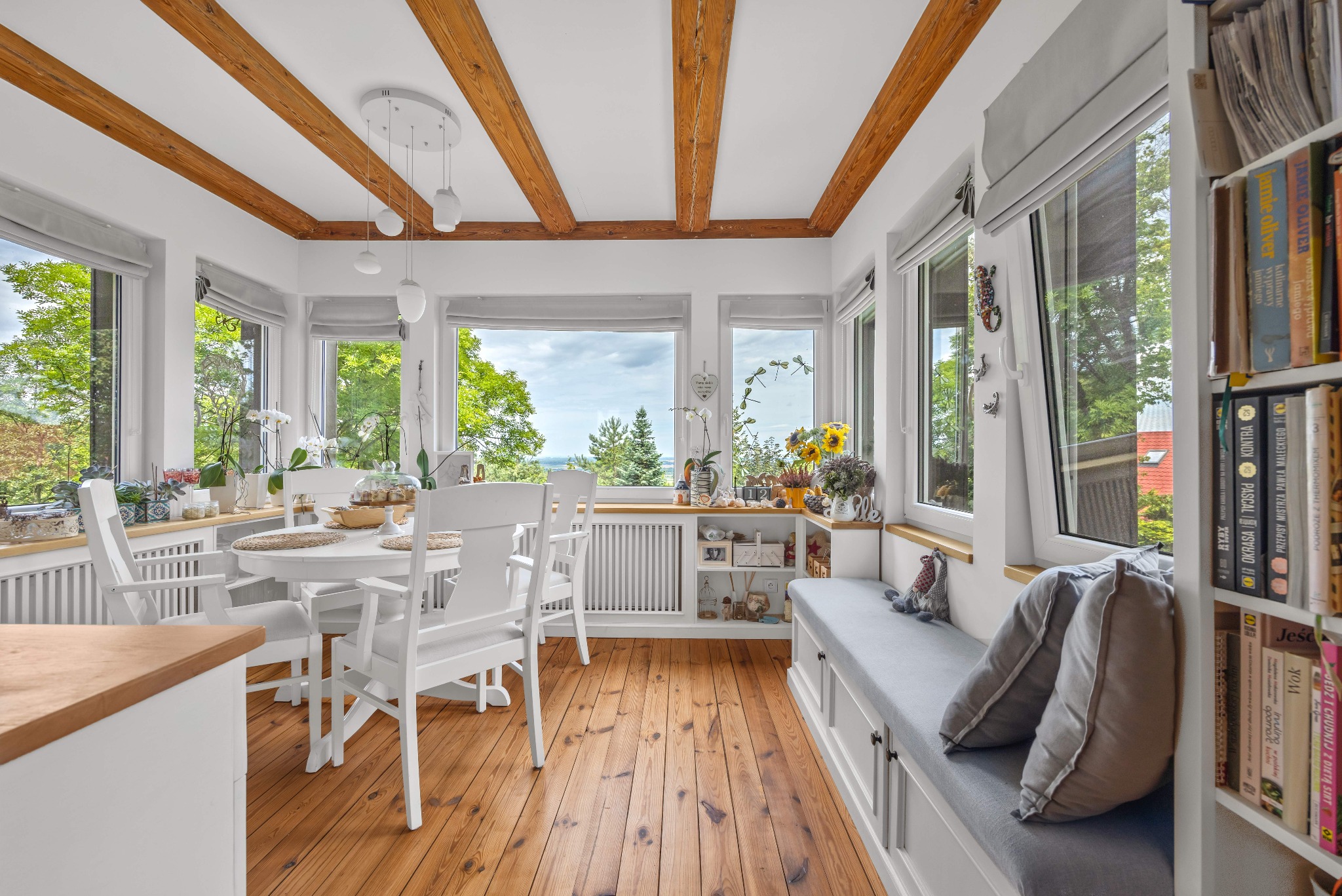 Jasne wnętrze jadalni z drewnianą podłogą i belkami stropowymi. Białe meble, duże okna z widokiem na zieleń. Przytulny kącik do czytania z poduszkami.