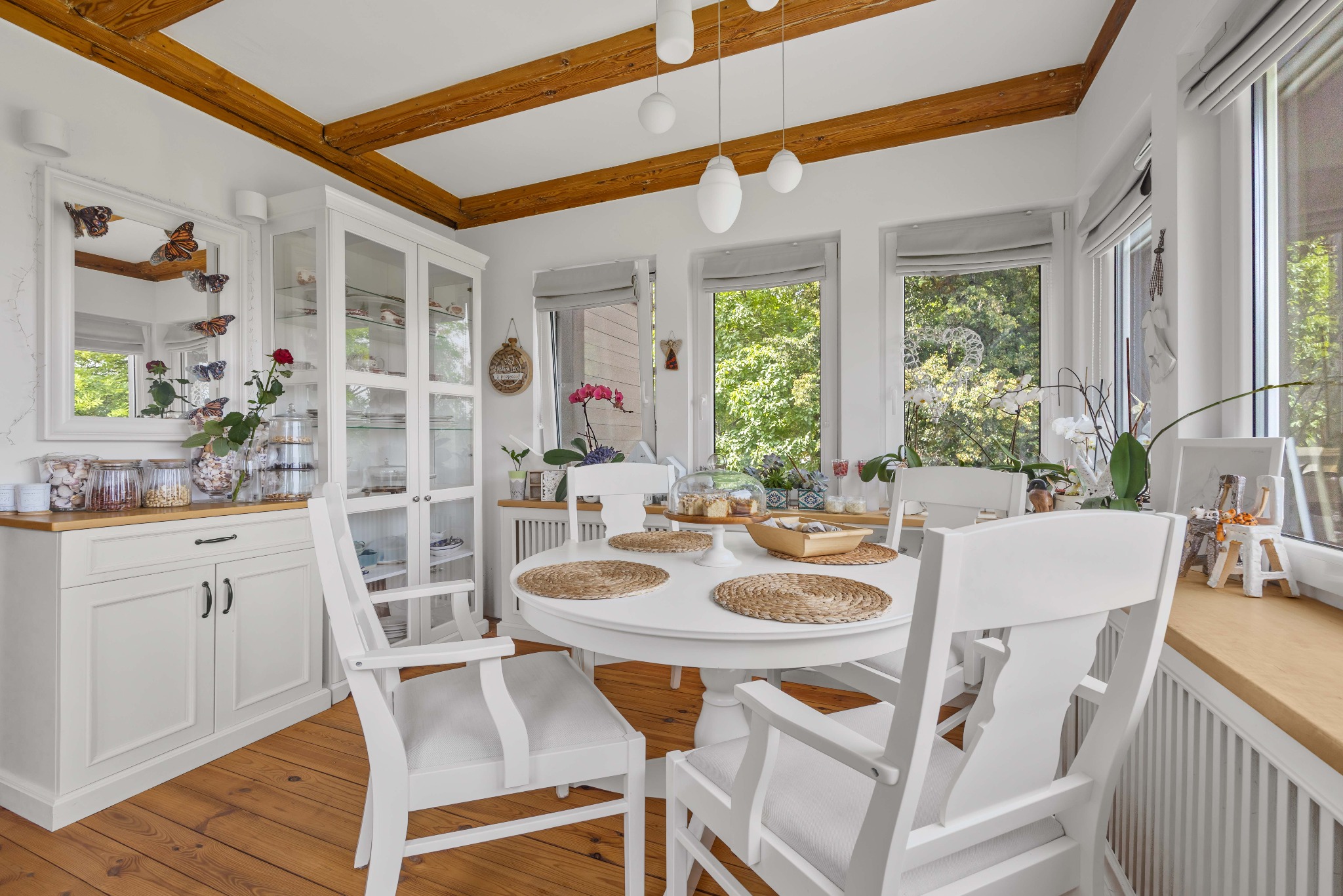 Jasna jadalnia z białym okrągłym stołem i krzesłami. Drewniane belki stropowe, przeszklona witryna i dekoracje z motywem motyli dodają wnętrzu unikalnego charakteru.