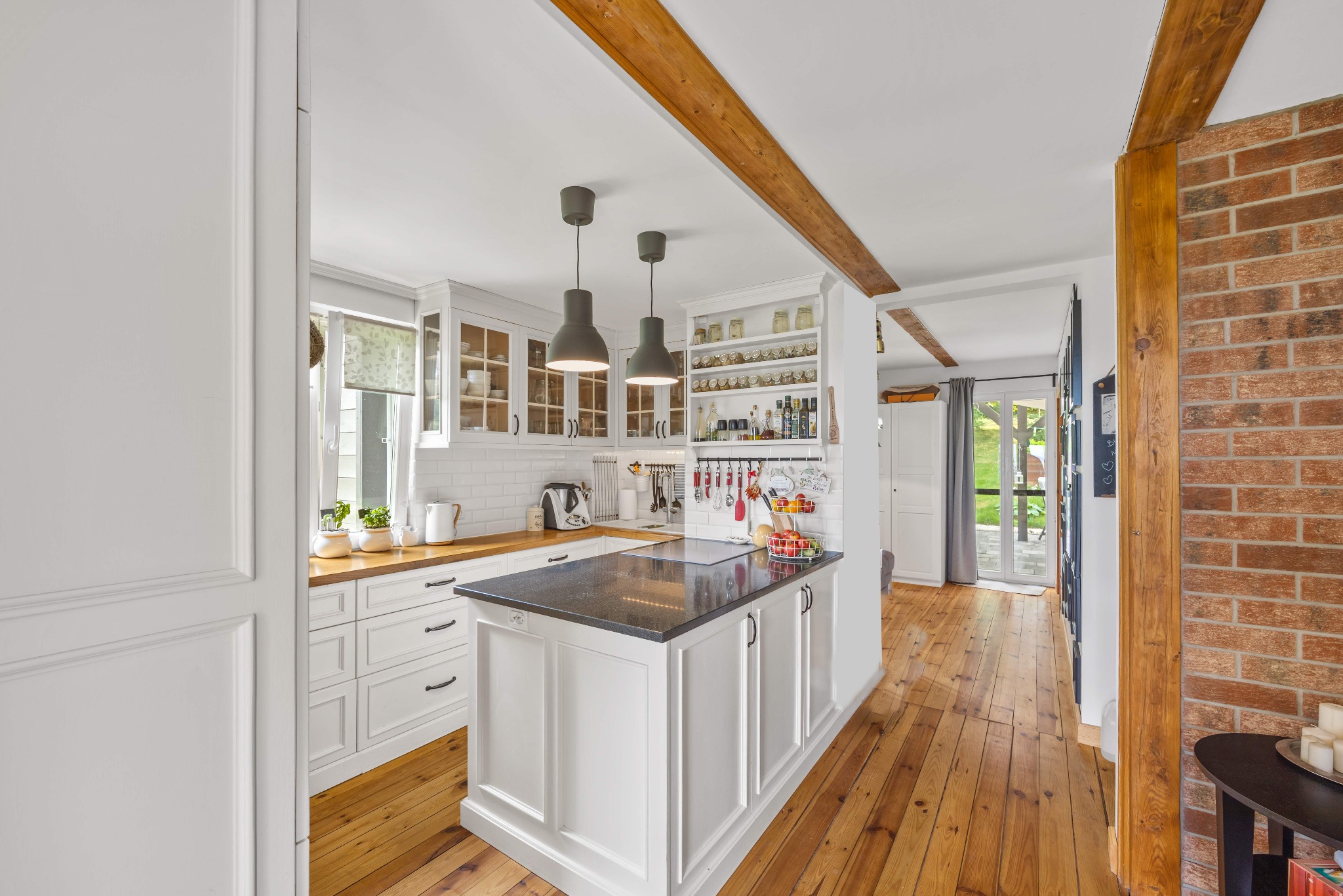Jasna kuchnia z wyspą i drewnianymi belkami stropowymi. Widok na okno i drzwi balkonowe. Parkiet z desek. Ceglana ściana. Dwie wiszące lampy.