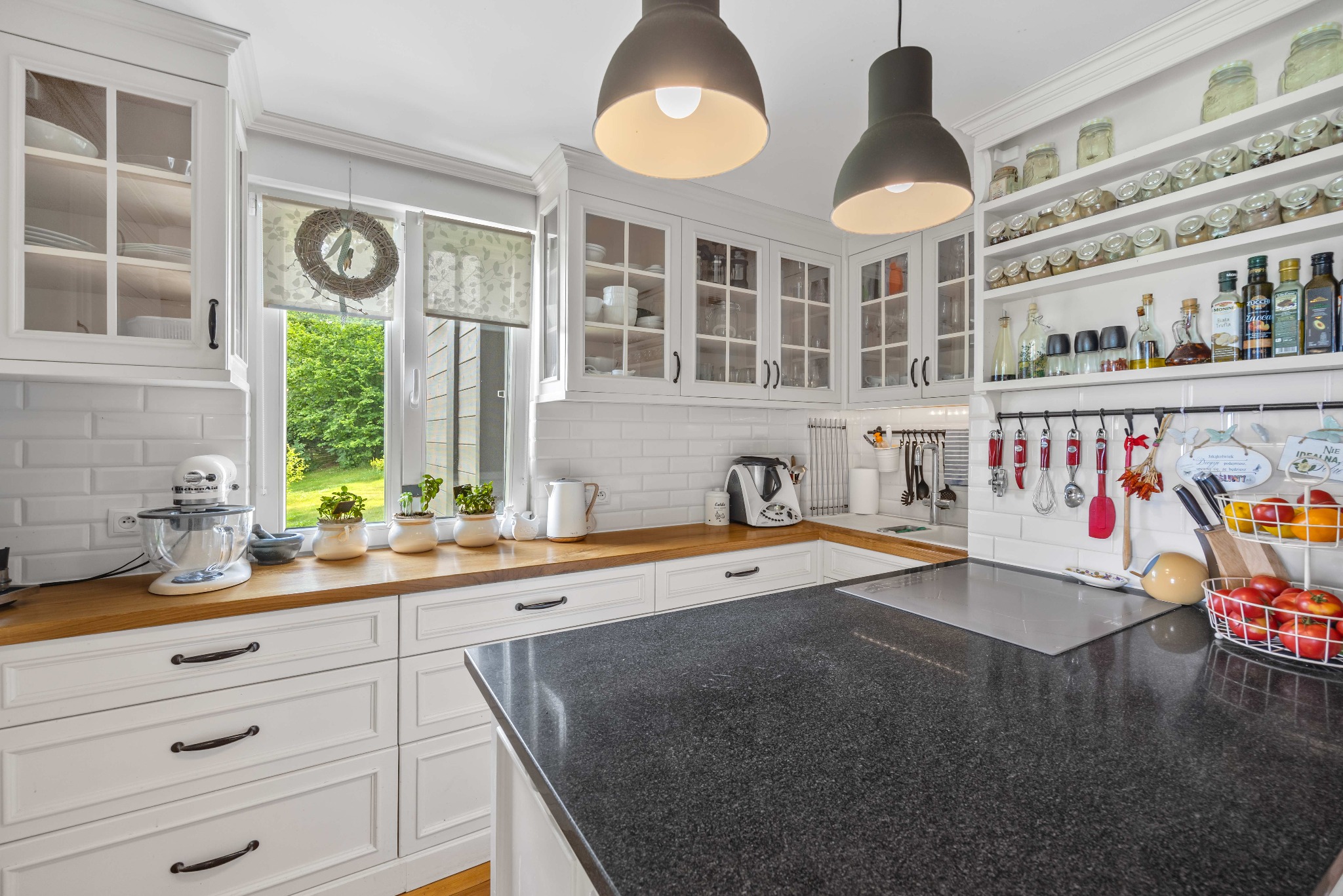 Jasna kuchnia w stylu rustykalnym: białe szafki z przeszklonymi frontami, drewniany blat, czarny kamienny półwysep, okno z wiankiem i widokiem na zieleń.