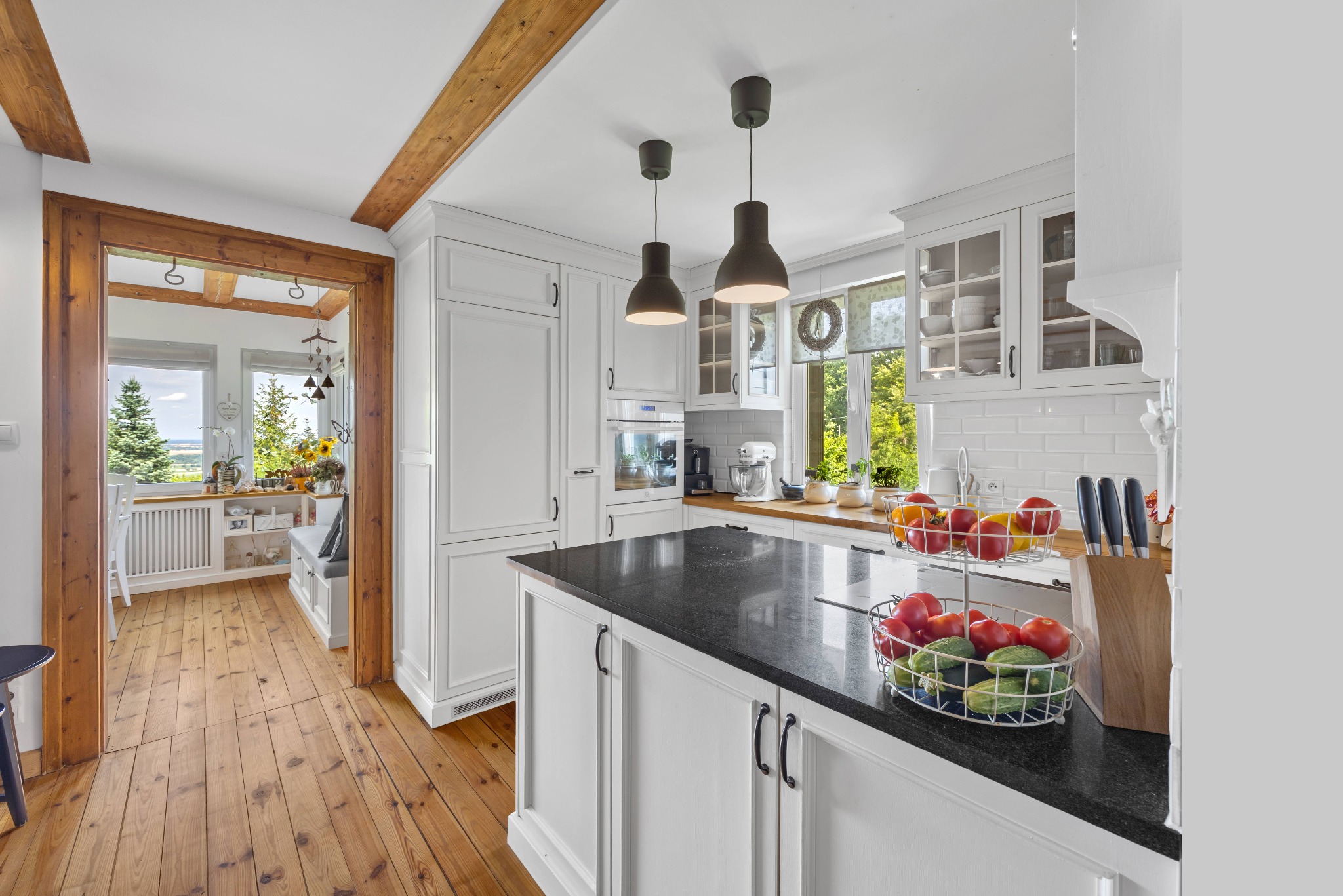 Jasna kuchnia w stylu rustykalnym: białe meble, czarny blat wyspy, drewniane belki stropowe i podłoga. Widok na jadalnię z oknem na zewnątrz.