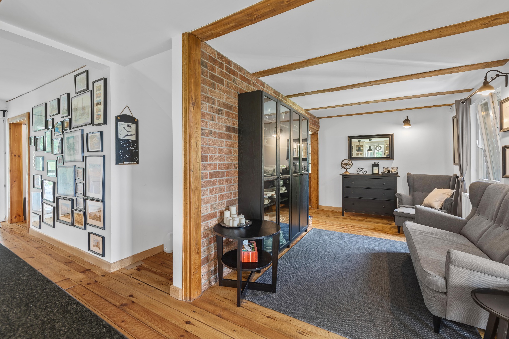 Jasne wnętrze z drewnianą podłogą, ceglaną ścianą i szarą sofą. Na ścianie galeria obrazów, a w głębi pokoju komoda i lustro. Widoczne drewniane belki stropowe.