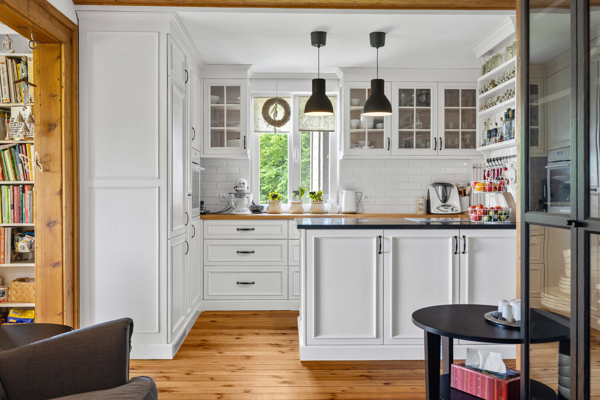 Jasna kuchnia w stylu klasycznym z białymi szafkami, drewnianym blatem i podłogą. Widoczne detale: okno z wiankiem, czarne lampy, książki na półce w tle.