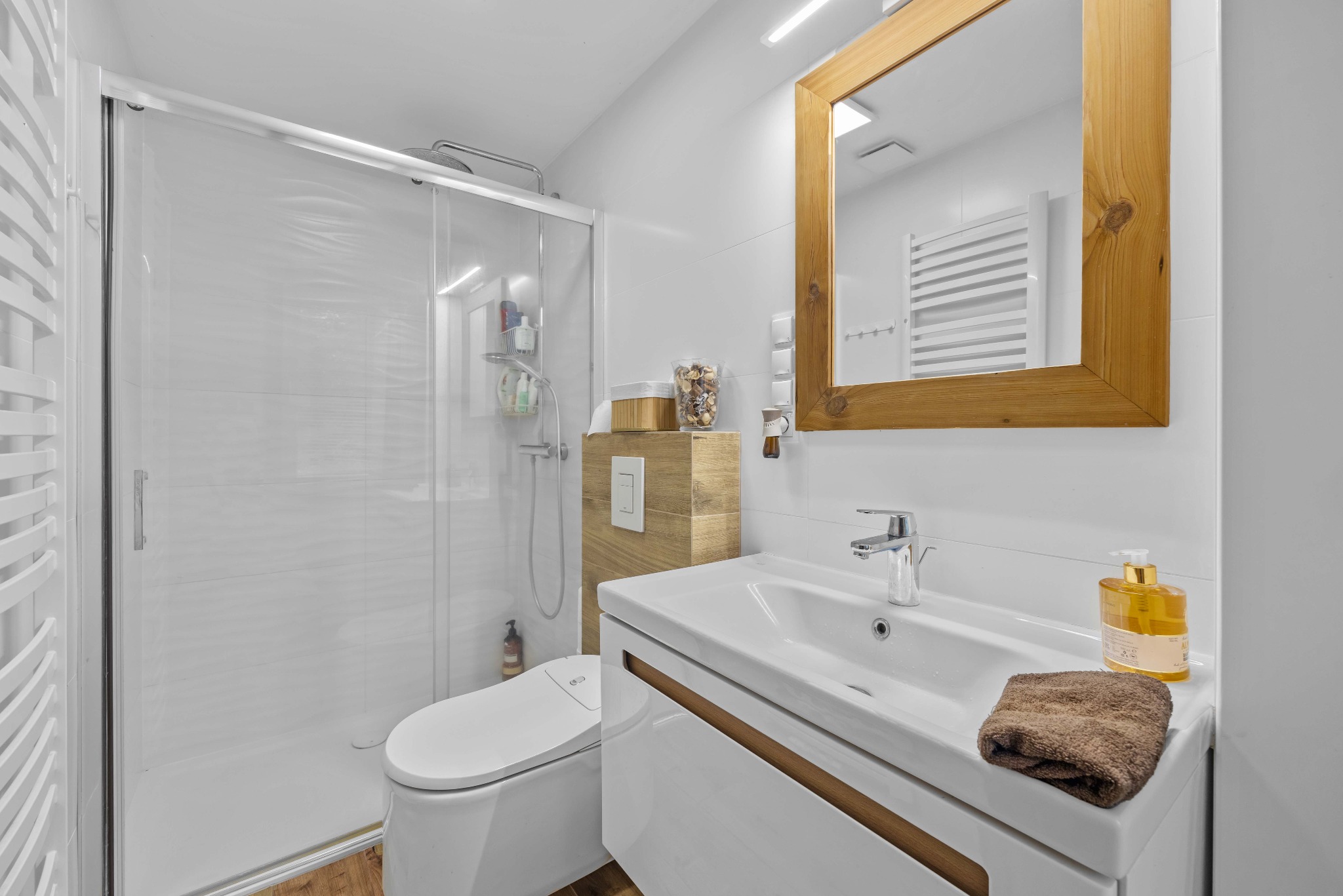 Nowoczesna łazienka z kabiną prysznicową, umywalką z szafką i lustrem w drewnianej ramie. Białe płytki, drewniane akcenty i brązowy ręcznik.