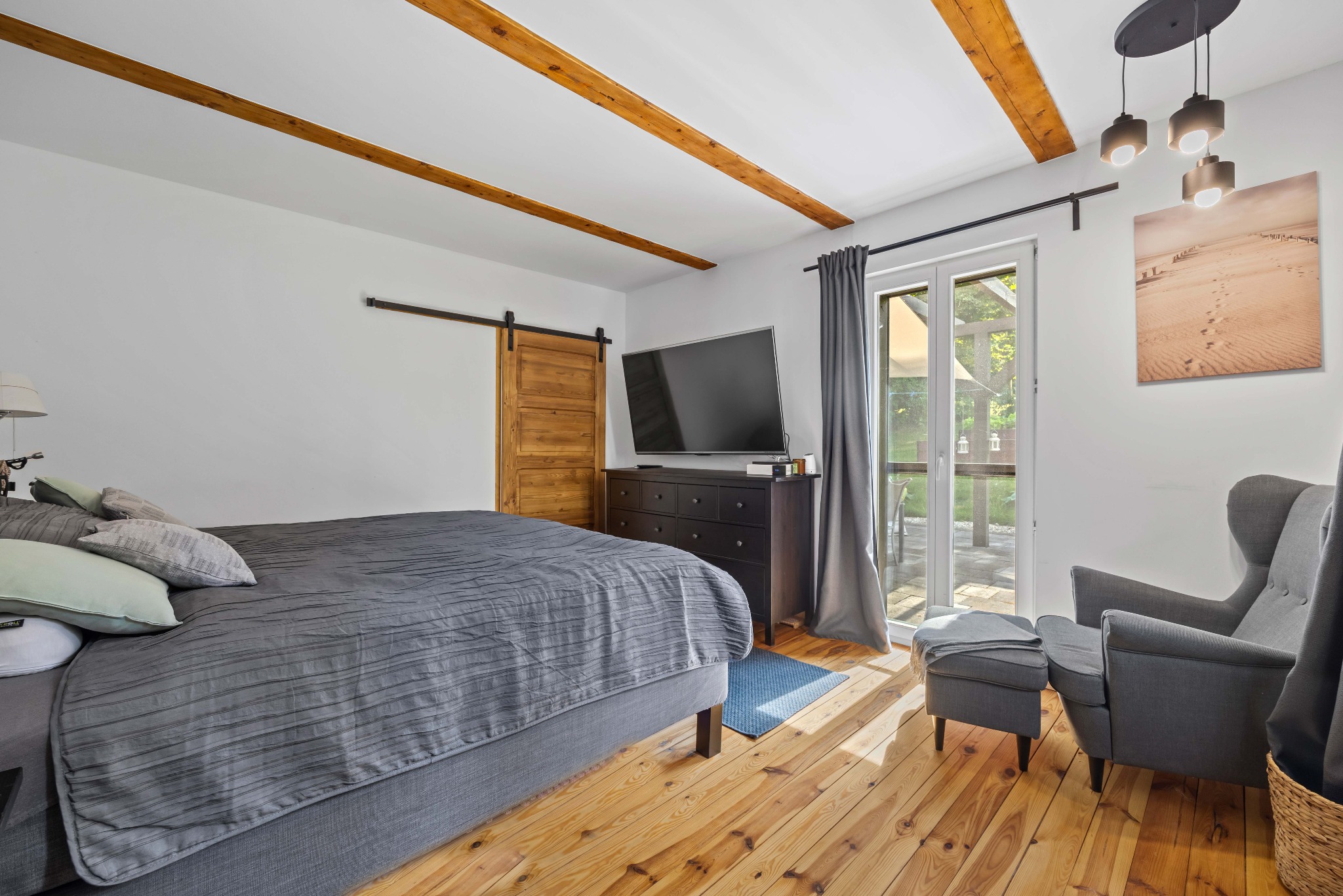 Jasna sypialnia z drewnianą podłogą i belkami stropowymi. Widoczne łóżko z szarą pościelą, komoda z telewizorem, fotel z podnóżkiem i drzwi przesuwne.