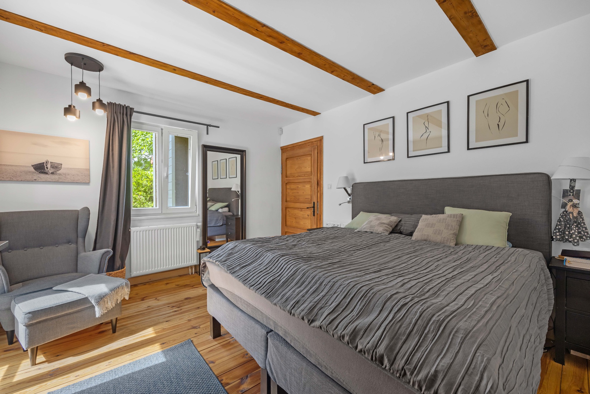 Jasna sypialnia z drewnianymi belkami stropowymi, szarym łóżkiem i fotelami. Na ścianach grafiki w czarnych ramkach, na podłodze drewniana podłoga i niebieski dywanik.