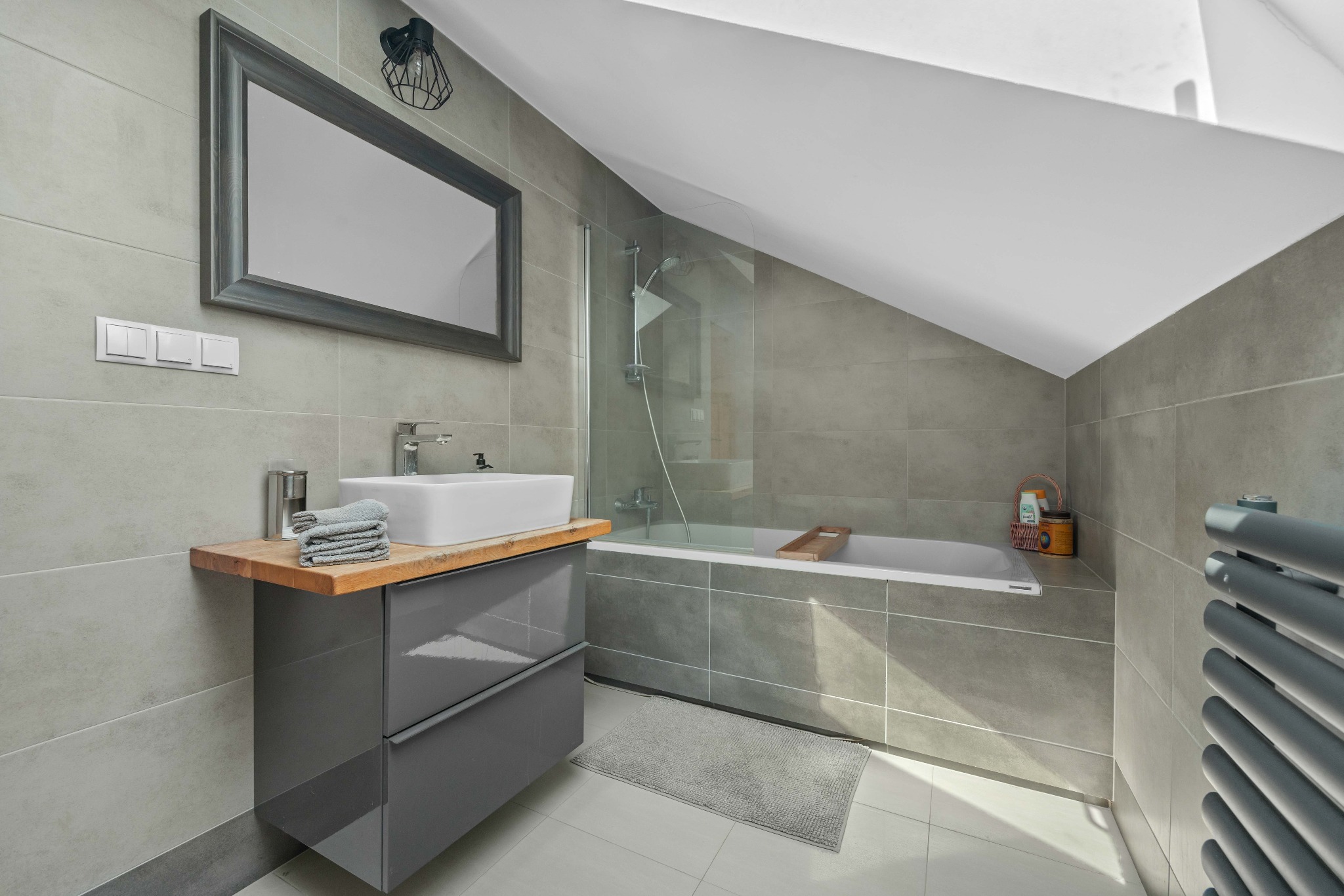 Jasna łazienka na poddaszu z wanną, umywalką na szafce z drewnianym blatem, lustrem w ramie. Szare płytki na ścianach i podłodze. Nowoczesny design.