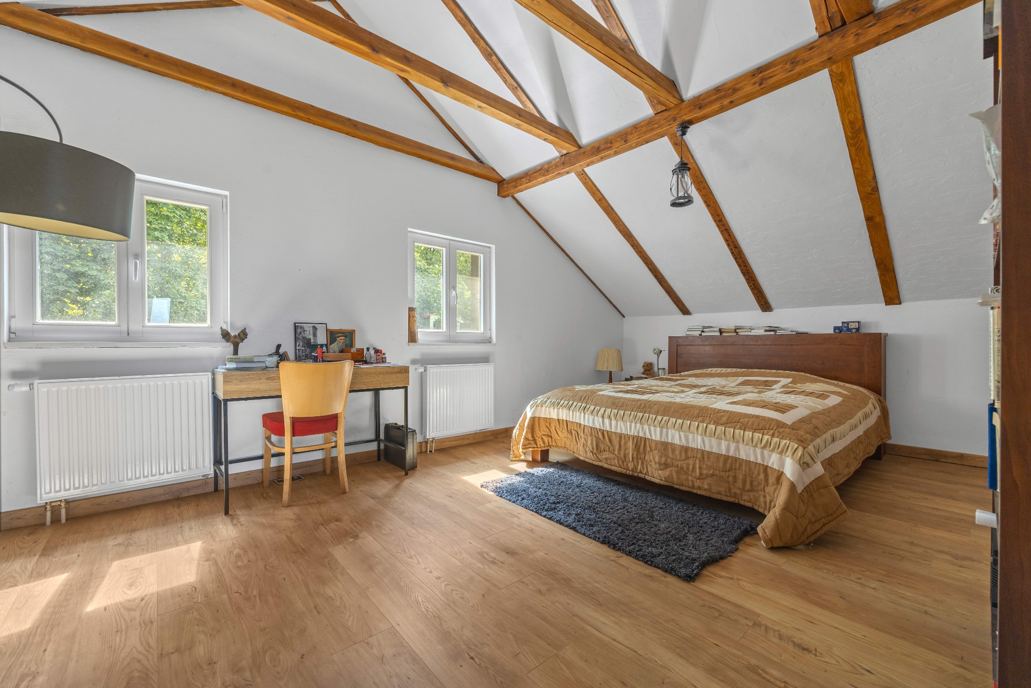 Jasne poddasze z drewnianą konstrukcją dachu, biurkiem przy oknie i łóżkiem z beżową narzutą. Wystrój uzupełnia dywanik i lampa wisząca.