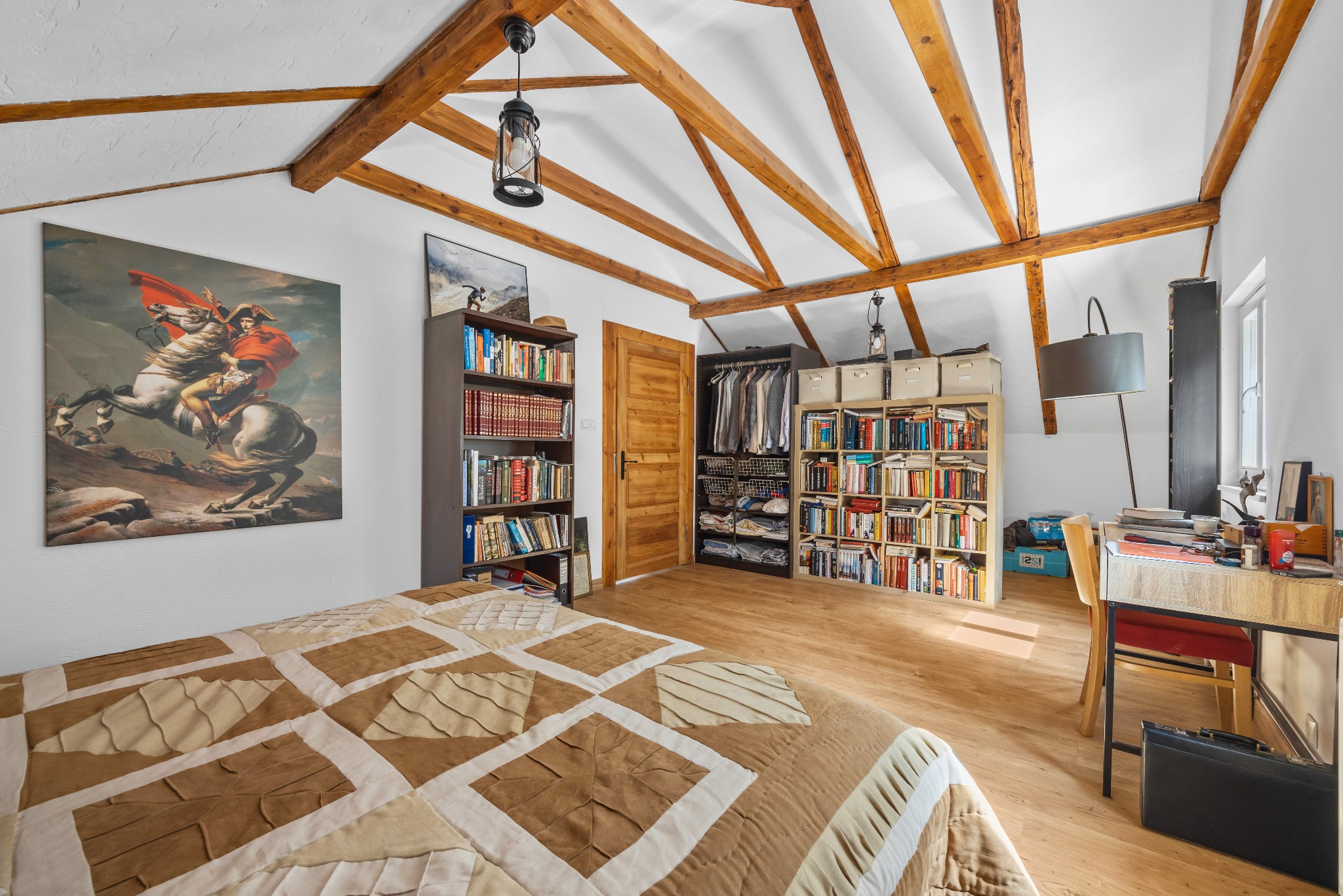 Jasna sypialnia na poddaszu z drewnianymi belkami, łóżkiem z patchworkową narzutą i biurkiem. Obraz Napoleona na ścianie dodaje charakteru.