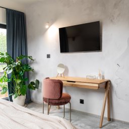 BARLOW SPÓŁKA Z OGRANICZONĄ ODPOWIEDZIALNOŚCIĄ - Nowoczesna sypialnia z biurkiem z jasnego drewna, różowym krzesłem, telewizorem na ścianie z artystycznym malunkiem, dużą rośliną doniczkową przy oknie i beżową narzutą na łóżku.