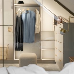 BARLOW SPÓŁKA Z OGRANICZONĄ ODPOWIEDZIALNOŚCIĄ - Wnętrze garderoby na poddaszu ze skośnym sufitem, oświetlenie LED wewnątrz, ubrania wiszące na wieszakach, pufa obita jasną tkaniną, widok z łóżka.