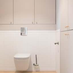 BARLOW SPÓŁKA Z OGRANICZONĄ ODPOWIEDZIALNOŚCIĄ - Nowoczesna łazienka z podwieszaną toaletą, białą boazerią ścienną, beżowymi szafkami z okrągłymi złotymi uchwytami i drewnianą podłogą.