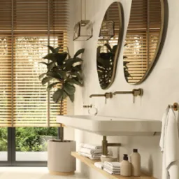 Nowoczesna łazienka z owalnymi lustrami w złotych ramach, minimalistyczną białą umywalką, drewnianymi żaluzjami w oknie i rośliną doniczkową, oświetlona przez szklaną lampę wiszącą.