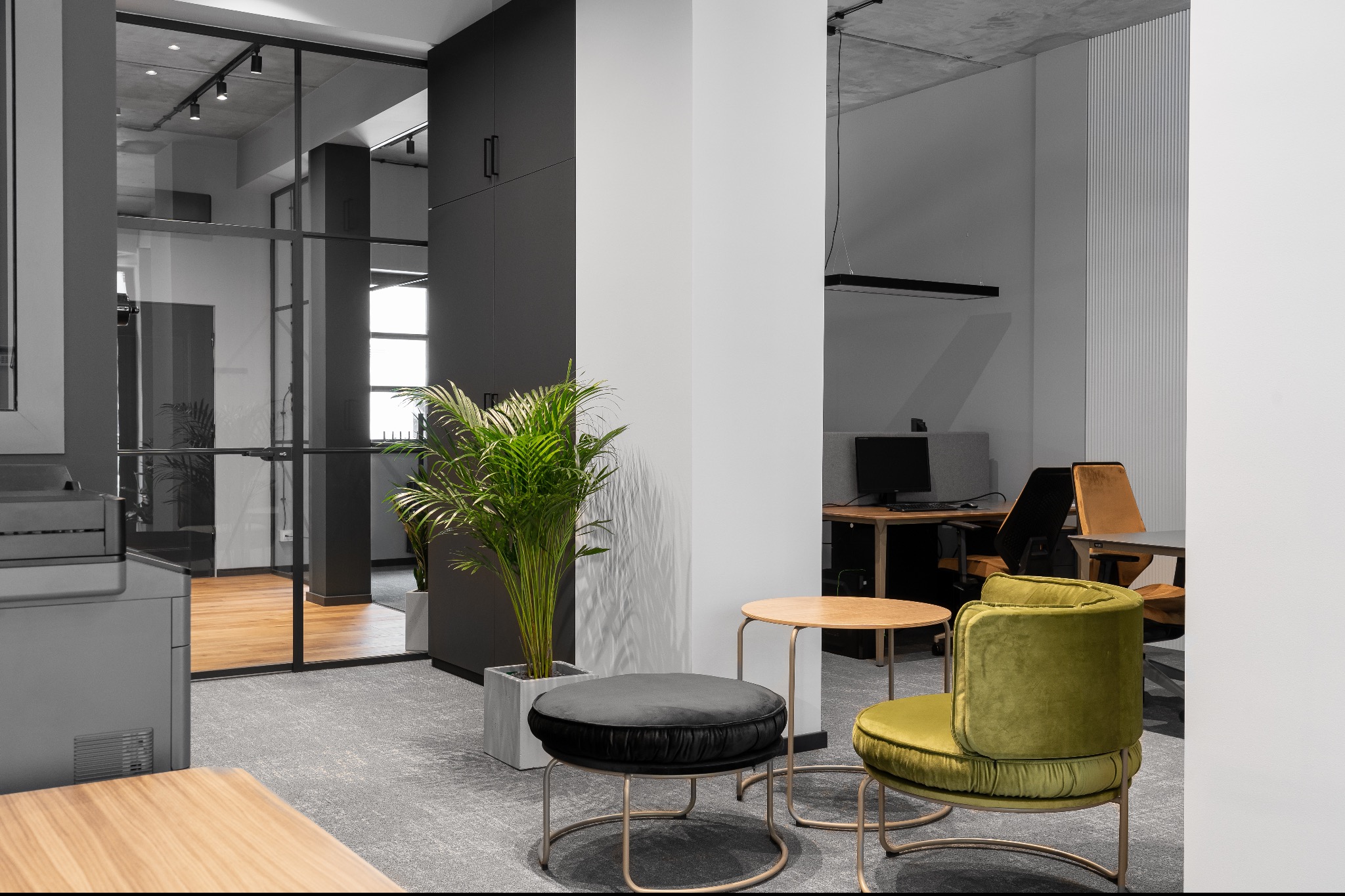 Nowoczesne biuro z przeszkloną ścianą działową, czarnymi szafami, rośliną doniczkową w betonowej donicy, okrągłym pufem, stolikiem kawowym i zielonym krzesłem na metalowych nogach, widok...
