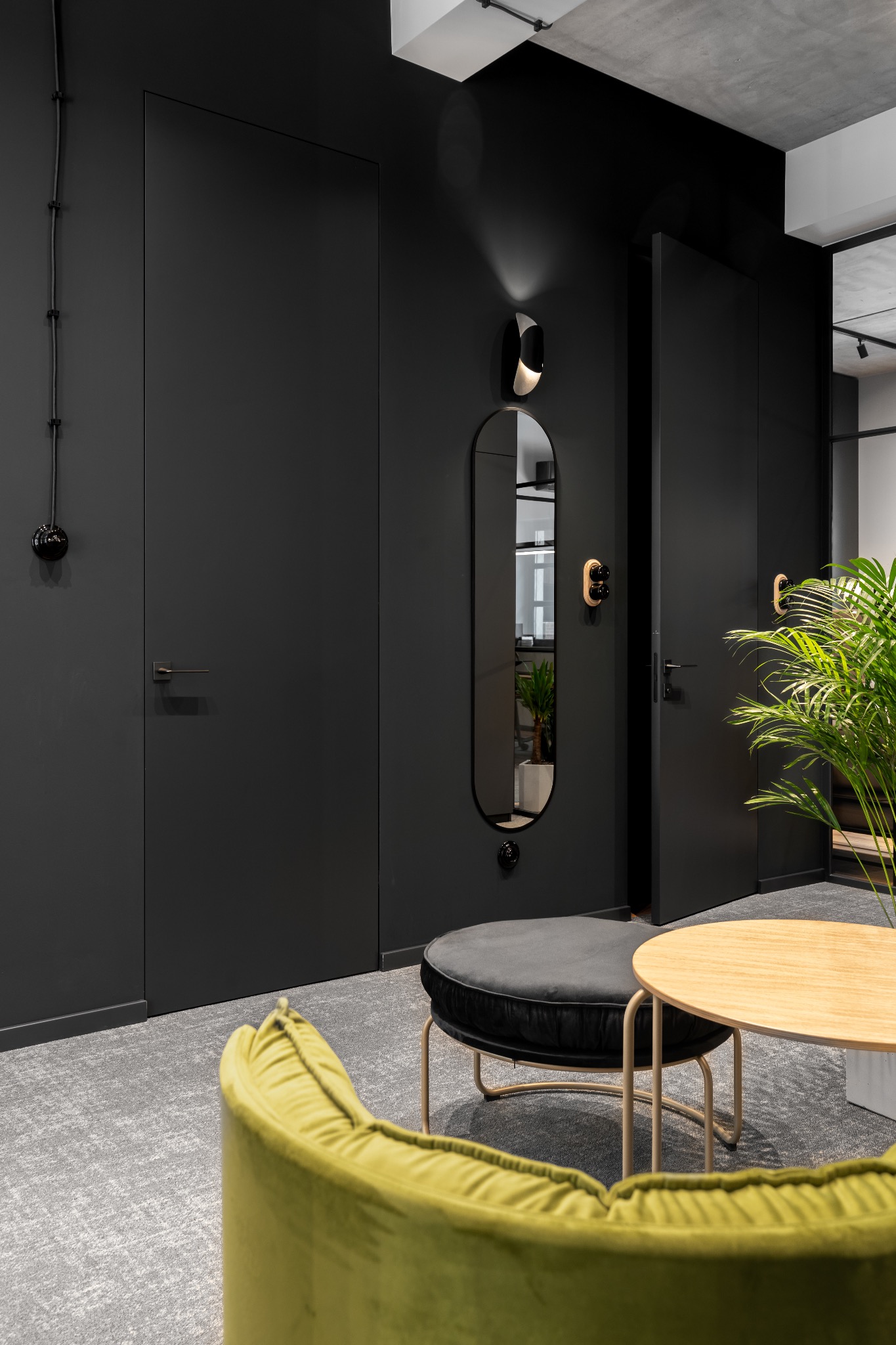 Nowoczesne wnętrze biurowe z czarnymi ścianami, ukrytymi drzwiami, owalnym lustrem z dekoracyjnym oświetleniem, zielonym fotelem, okrągłym stolikiem i rośliną doniczkową.