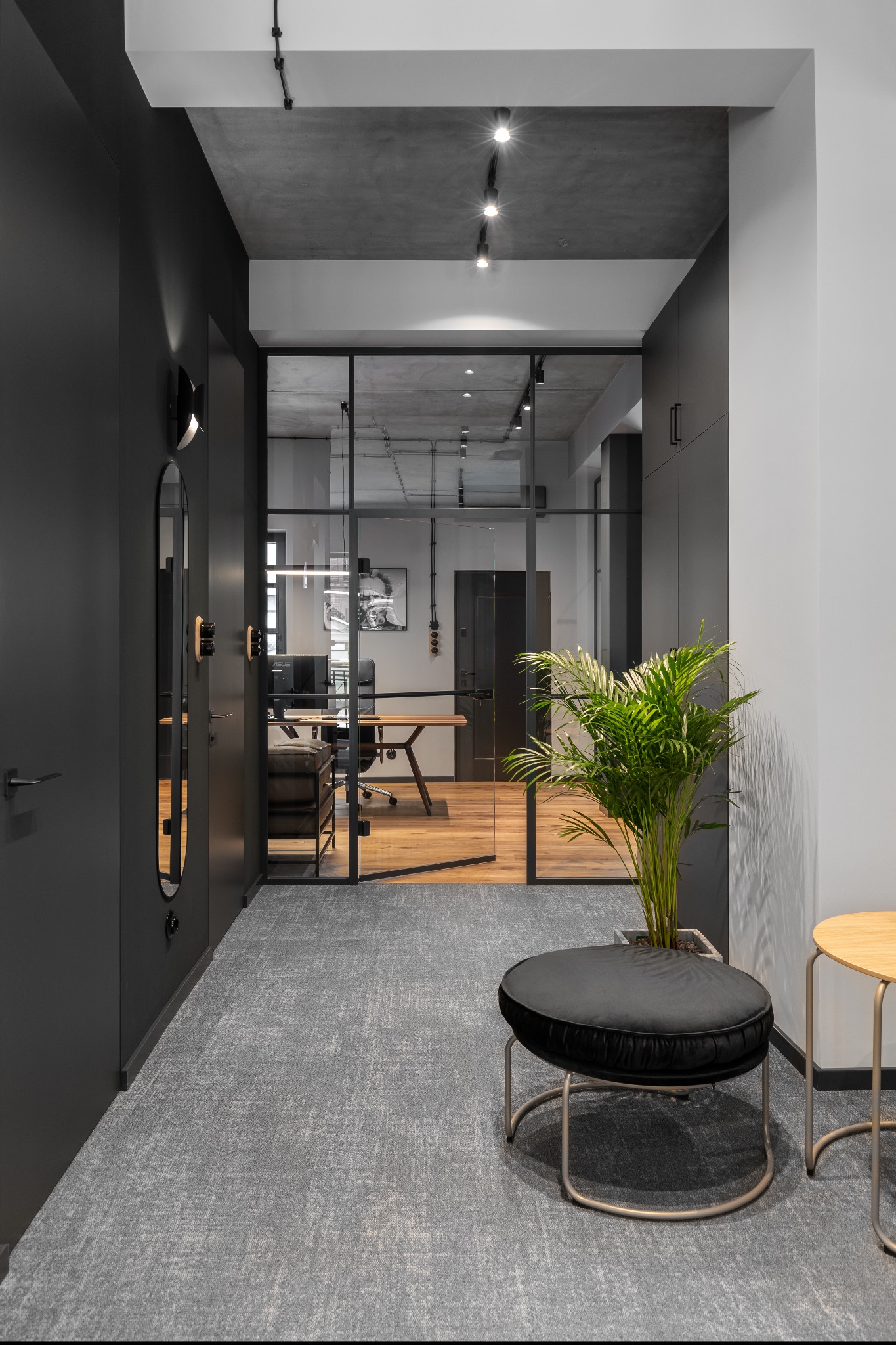 Nowoczesne biuro z betonowym sufitem, szklaną ścianą działową, drewnianym biurkiem, czarnymi ścianami, pufem i rośliną doniczkową w minimalistycznym stylu.