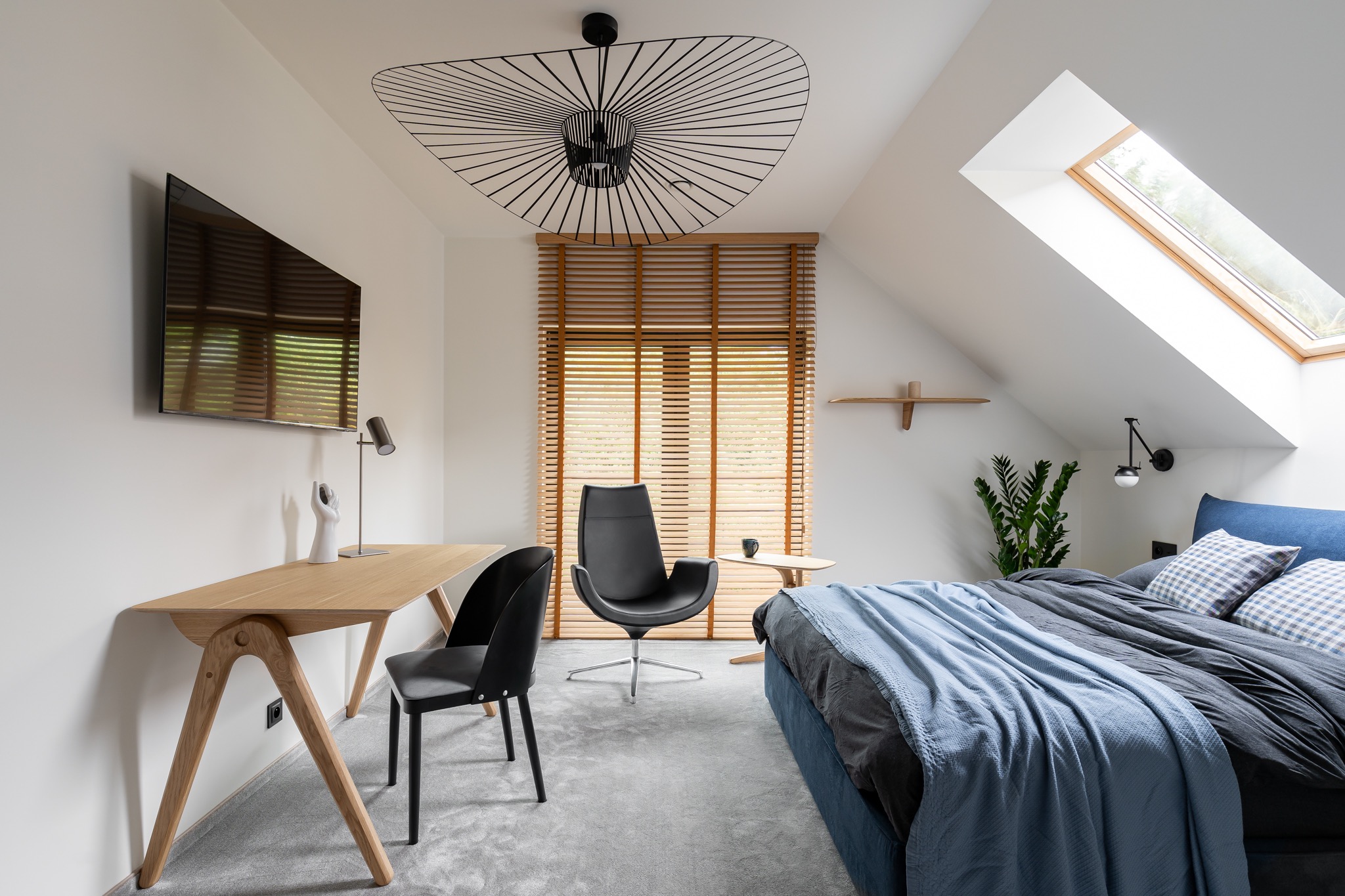 Pokój na poddaszu z drewnianym biurkiem, krzesłami, łóżkiem z niebieską narzutą i oryginalną lampą sufitową w kształcie wachlarza.