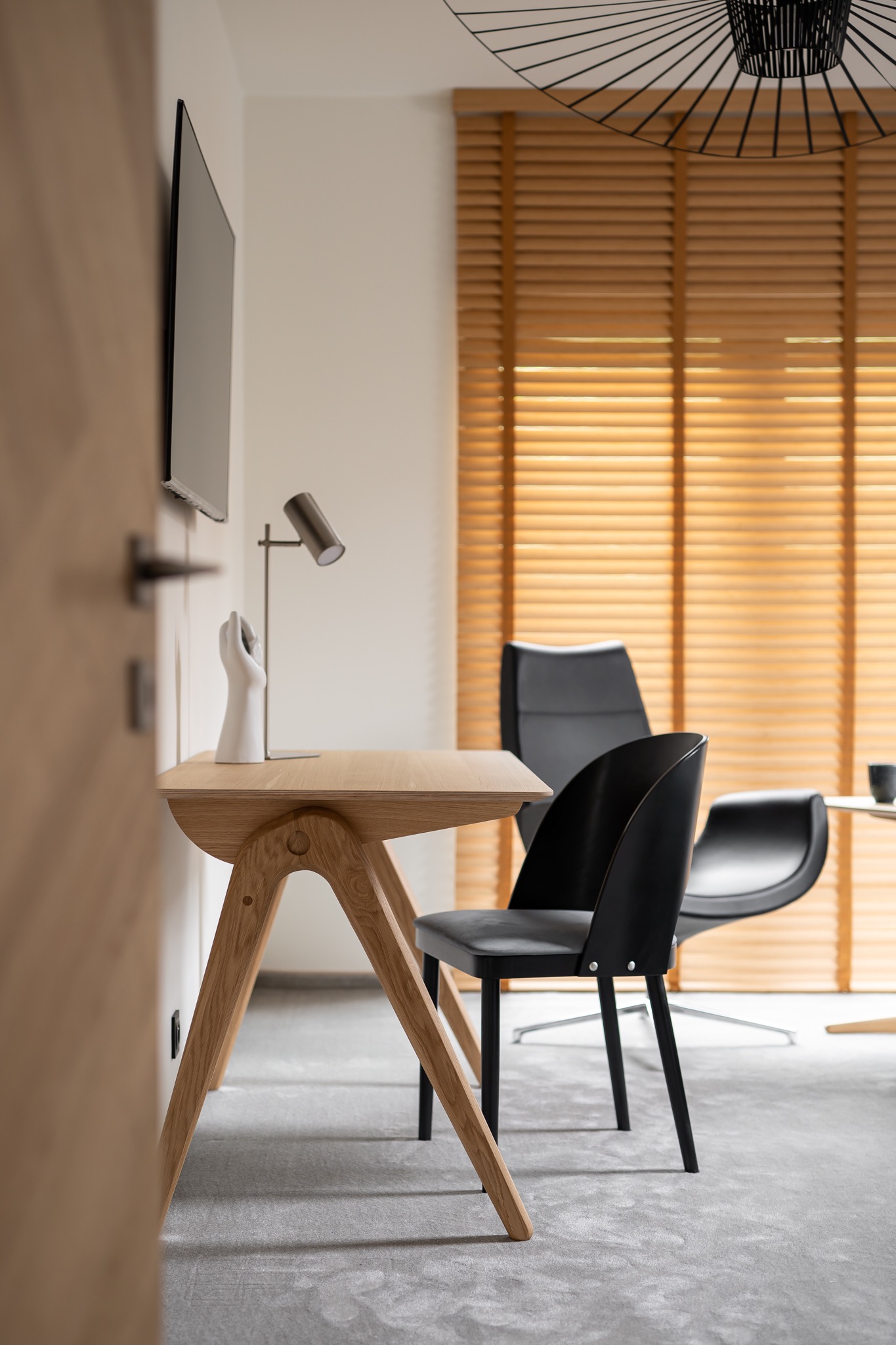 Nowoczesne biuro domowe z drewnianym biurkiem o unikalnej konstrukcji nóg, czarnym krzesłem, lampką biurkową i ceramiczną rzeźbą dłoni na tle żaluzji okiennych.